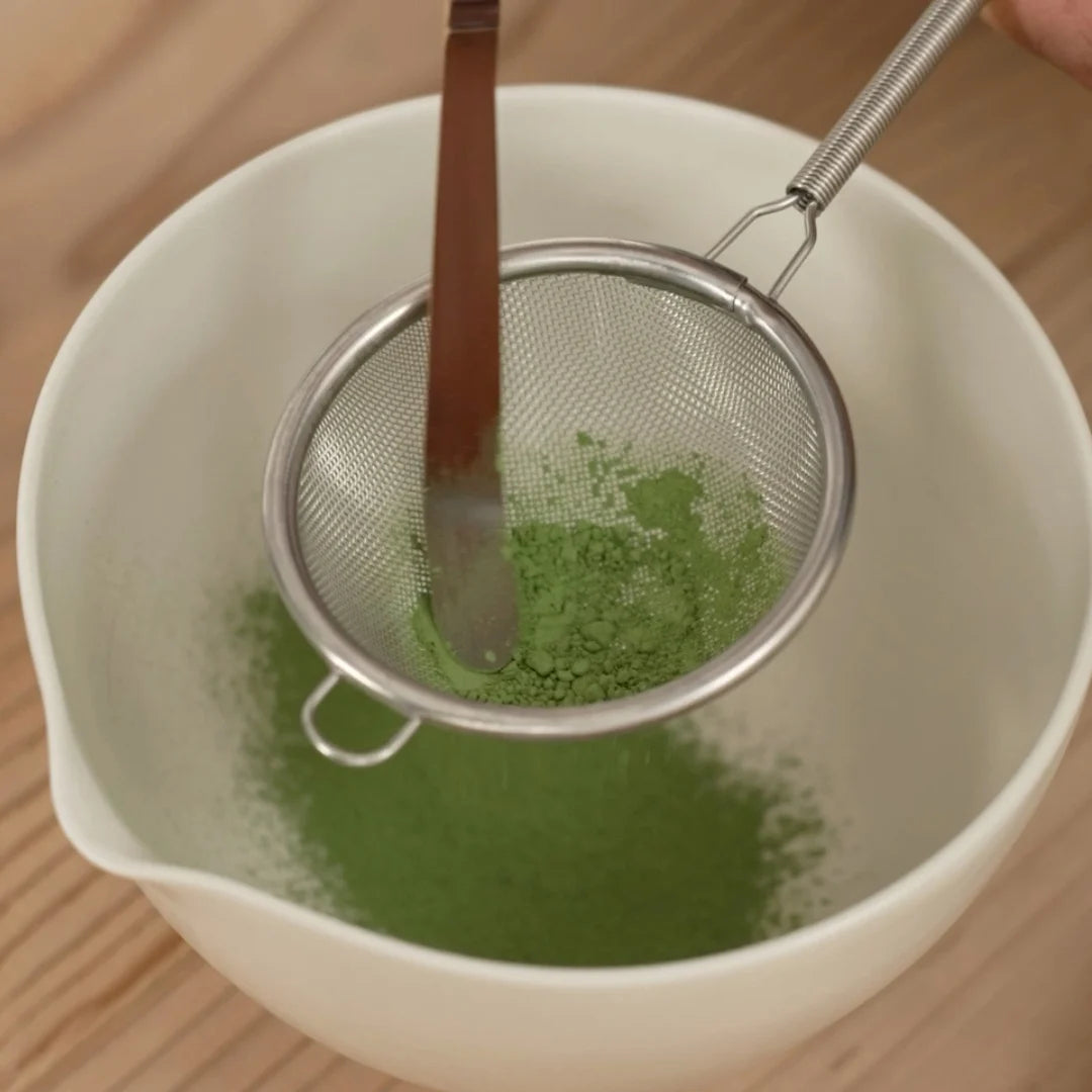 Green powder being sifted into a white bowl with a metal sieve.