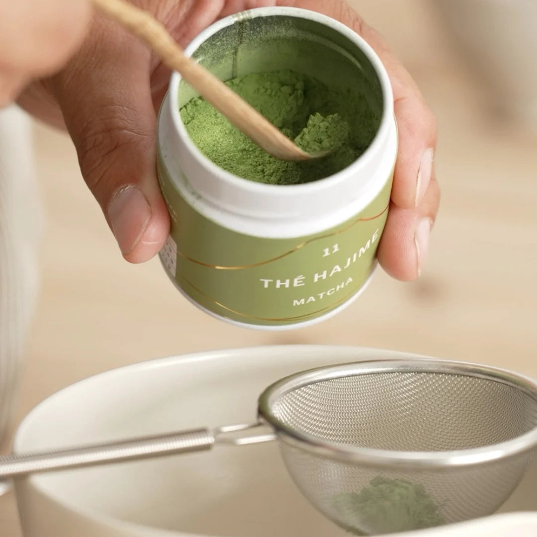 Person holding a container of matcha green tea powder with a bamboo spoon and a metal sieve on a light background.