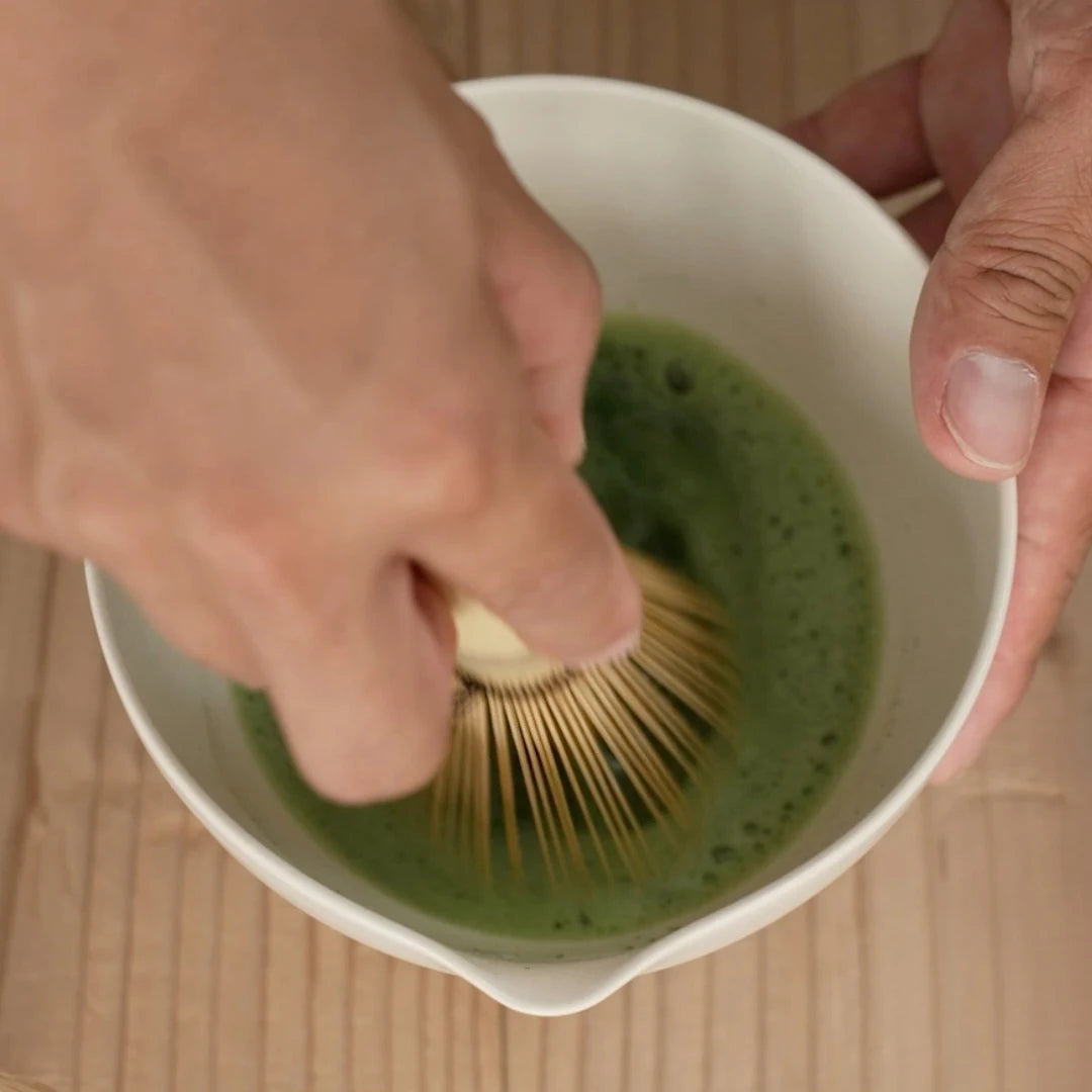 A person using a chasen to whisk matcha in a white bowl on a wooden surface