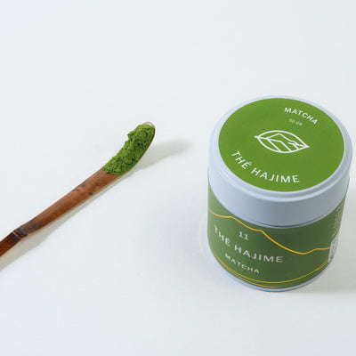A bamboo spoon (Chasaku) next to a white and green labeled Matcha powder tin can on a white background.